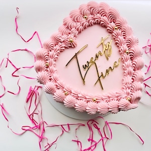 May include: Pink heart-shaped cake with gold lettering that says "Twenty Nine" on top. The cake is decorated with pink frosting and gold sprinkles.