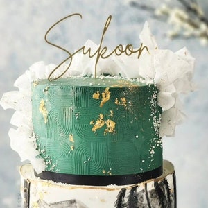May include: A two-tiered cake decorated with a green geometric pattern on the top tier and a black and white ink painting of a mountain landscape on the bottom tier. The cake is topped with a gold topper that says "Sukoon".