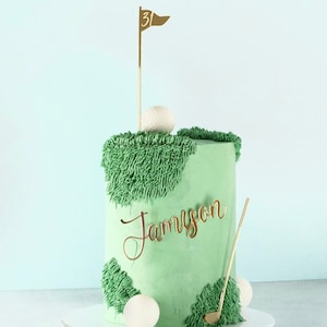 May include: A light green cake decorated to resemble a golf course. The cake has green frosting, with textured green frosting accents. Gold accents include a golf flag, golf club, and the name "Jamyson". White golf balls are also included.