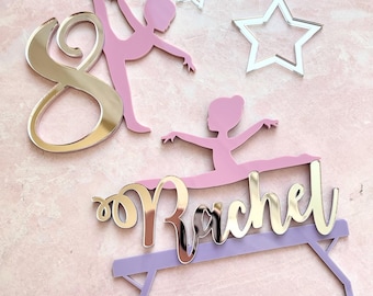 May include: A collection of ballet-themed cake toppers and decorations. Includes a mirrored gold number 8, pink ballerina figures, silver stars, and a topper with the name "Rachel" in gold. The pieces are set against a pink marble background.