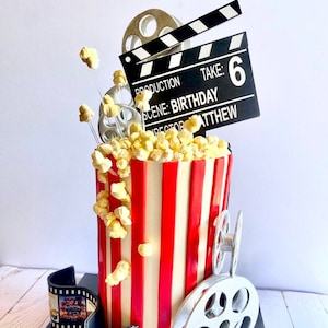 May include: A red and white striped popcorn bucket cake decorated with movie themed decorations, including a film reel, clapperboard, and popcorn. The clapperboard reads "Production, Scene: Birthday, Take: 6, Director: Matthew".