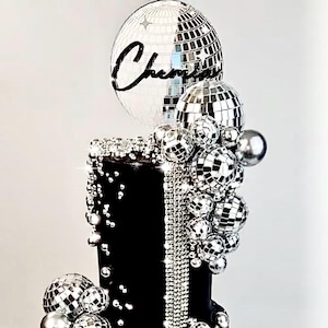 May include: A black cake decorated with silver disco balls and rhinestones. The cake is topped with a large disco ball and the word "Cheers" in black script. The cake has a glamorous, celebratory theme.