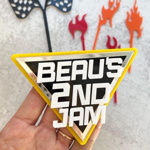 May include: A yellow, black, and silver cake topper with the text "BEAU'S 2ND JAM" in white. The topper is in the shape of a triangle. Additional toppers include checkered flags and flames.