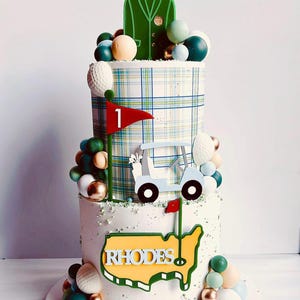 May include: A two-tiered cake decorated with a golf theme. The bottom tier is white with a green and yellow outline of the United States and the word "RHODES" in yellow letters. The top tier is covered in a plaid pattern with a green golf cart and a red flag with the number "1" on it. The cake is surrounded by small, colorful balls.