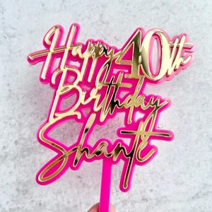 May include: A cake topper featuring a bright pink acrylic base with a gold mirrored acrylic overlay. The text reads Happy 40th Birthday Shante in a cursive and print font combination. A single pink stake extends from the bottom for insertion.