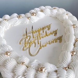 May include: A white heart-shaped cake decorated with white frosting and gold sprinkles. The cake is topped with a gold glitter topper that says "Happy Birthday Chris".