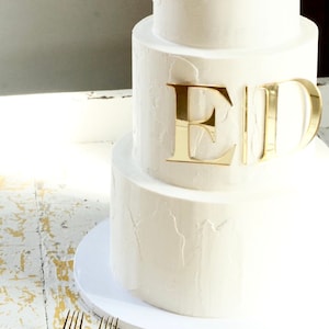 May include: A three-tiered white wedding cake with gold foil letters spelling "ED" on the top tier.