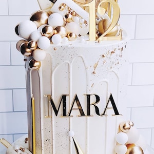 May include: A white cake with gold accents, featuring the name "MARA" in gold lettering. The cake is decorated with gold and white spheres, chocolate bars, and a "12" cake topper. Gold candles and a cat figurine complete the festive design.