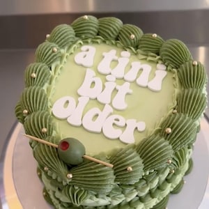 May include: A square-shaped cake with light green frosting and decorative green frosting swirls around the edges. The cake has the words "a tini bit older" in white lettering. An olive on a toothpick is placed on the cake, and gold sprinkles are added.