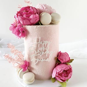 May include: A pink frosted birthday cake decorated with pink flowers, macarons, and the words "Happy Birthday Emma" in white script. The cake is on a white cake stand.