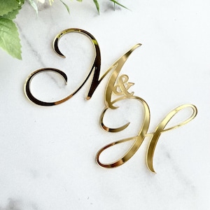 May include: A gold-colored wedding cake topper featuring the intertwined letters "M" and "H", with an ampersand symbol between them. The elegant, cursive design is set against a white marble background, with a hint of greenery and pink flowers.
