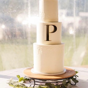 May include: A three-tiered white wedding cake with delicate floral designs. The top tier has a gold letter "P" on it. The cake is on a wooden cake stand with a black metal base.