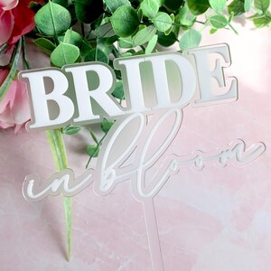 May include: White acrylic cake topper with the text "Bride in Bloom" in a cursive font.
