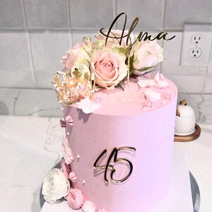 May include: A pink cake decorated with white flowers, a gold crown, and gold lettering that says "Alma" and "45".