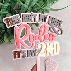 Könnte beinhalten: Ein rosa-weißer Tortenaufsatz mit dem Text "This ain't my first Rodeo, It's my 2nd". Der Aufsatz hat ein mehrschichtiges Design mit einer rosa Basis und verschiedenen Rosa-, Grau- und Weißtönen. Ein kleiner rosa Stiefel ist sichtbar.