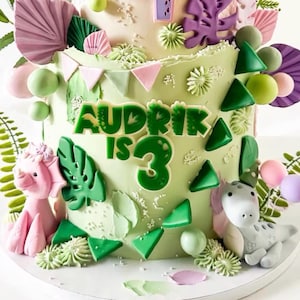 May include: A three-tiered birthday cake with green frosting, decorated with dinosaur figures, green leaf accents, and the name "Audrik is 3" in green lettering. The cake features pink, green, and purple decorative elements.