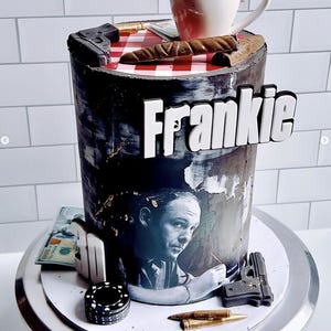 May include: A black and white cake decorated with a picture of a man, a red and white checkered tablecloth, a cigar, a gun, and the word "Frankie".