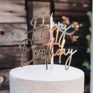May include: A gold-colored "Happy Birthday" cake topper with elegant cursive script, placed atop a white frosted cake. A white candle is positioned behind the topper. The background features a wooden surface.