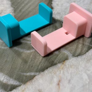 May include: Two 3D-printed holders, one teal and one pink, are displayed. Each has a rectangular base with two upright supports. The pink holder has a small square cutout. The objects are on a marbled surface.