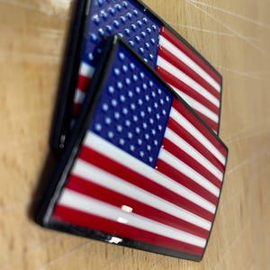 May include: Two rectangular enamel pins featuring the United States flag design. Each pin displays a blue canton with white stars in the upper left corner, followed by alternating horizontal red and white stripes. The pins have a glossy finish.