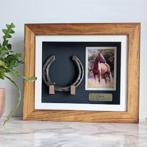 Könnte beinhalten: Gerahmtes Andenken mit einem Hufeisen und einem Foto eines braunen Pferdes. Der Holzrahmen umgibt einen dunklen Hintergrund mit einem Hufeisen und einem Foto eines Pferdes. Ein kleines Schild lautet "Clara 2007-2021".