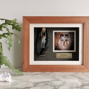 May include: A wooden shadow box memorial frame with a photo of a dog, a small glass vial with a paw print charm, and a leather dog collar. The plaque below the photo reads "Ranger 9/22/2009 - 3/1/2023".