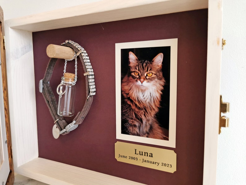 May include: A wooden shadow box memorial with a photo of a tabby cat named Luna. The box also includes a brown leather collar with a small glass vial and a silver chain with a charm.