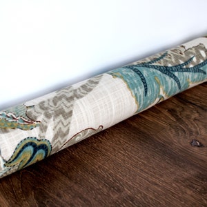 Op de afbeelding: Een beige deurstop met een blauw en bruin bloemenpatroon. De deurstop is opgerold en ligt op een houten vloer.