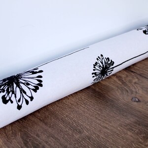 Op de afbeelding: Een witte deurstop met een zwarte paardenbloemprint. De deurstop is opgerold en ligt op een houten vloer.