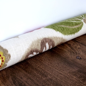 Op de afbeelding: Een witte deurstop met een bloemenpatroon in tinten groen, geel, rood en bruin. De deurstop is opgerold en geplaatst op een houten vloer.