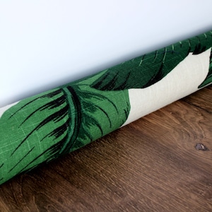 May include: Rolled-up fabric featuring a vibrant green and black tropical leaf pattern on a cream background. The fabric is positioned diagonally, showcasing the detailed leaf design. The background is a dark wood surface.