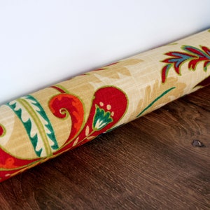 Op de afbeelding: Een beige deurstop met een kleurrijk bloemenpatroon in tinten rood, groen, blauw en geel. De deurstop is opgerold en ligt op een houten vloer.