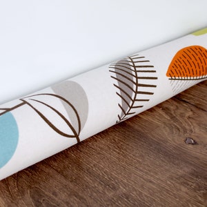 Op de afbeelding: Gerolde stof met een herhalend bladpatroon in oranje, grijs, blauw en groen op een witte achtergrond. De stof ligt op een houten oppervlak.