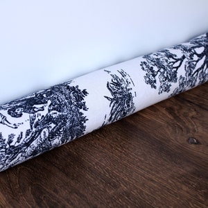 Puede incluir: Tela enrollada con un diseño clásico toile de Jouy en azul marino sobre fondo crema. El patrón representa escenas detalladas de árboles, figuras y paisajes. La tela está sobre una superficie de madera oscura.