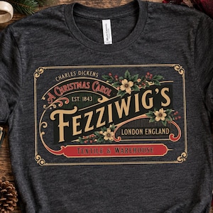 Pode incluir: Uma t-shirt cinzento-carvão com um gráfico retangular contendo o texto "Charles Dickens A Christmas Carol Est. 1843 Fezziwig's London England Textile & Warehouse" rodeado por ilustrações florais e de bagas em dourado, vermelho e verde.
