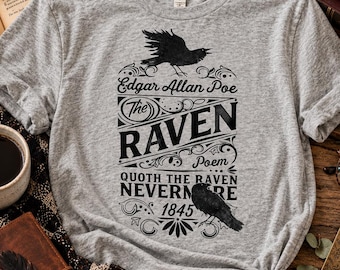May include: A heather gray t-shirt featuring a black graphic print of Edgar Allan Poe's The Raven. The design includes two raven illustrations, the text The Raven Poem, the quote Quoth the Raven Nevermore, and the year 1845 in a decorative font.