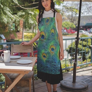 May include: A woman wearing a green and blue apron with a peacock feather design. The apron has a black strap and is tied at the waist. The woman is standing in front of a wooden table with two white plates and a white mug.