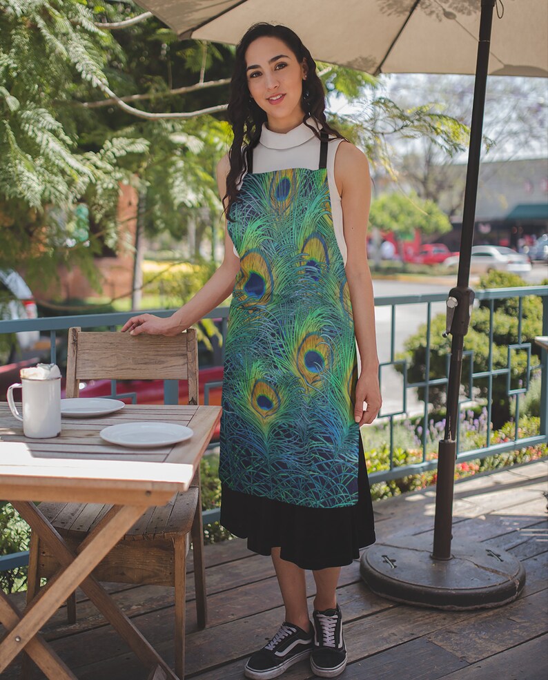 May include: A woman wearing a green and blue apron with a peacock feather design. The apron has a black strap and is tied at the waist. The woman is standing in front of a wooden table with two white plates and a white mug.