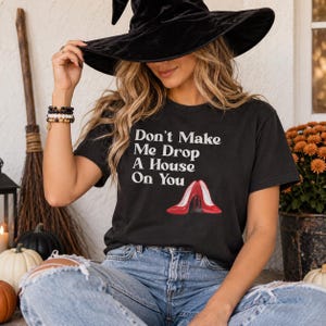 Pode incluir: Uma t-shirt preta de manga curta com o texto branco Don't Make Me Drop A House On You sobre um gráfico de um par de sapatos de salto alto vermelhos brilhantes. A t-shirt é usada com calças de ganga azuis e um chapéu de bruxa de veludo preto.