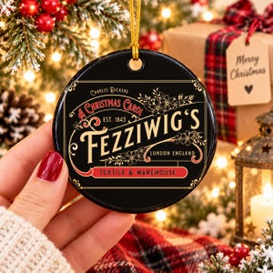 Pode incluir: Um enfeite de Natal redondo de cerâmica preta com texto em dourado e vermelho que diz Charles Dickens A Christmas Carol Est. 1843 Fezziwig's London England Textile & Warehouse, decorado com arte floral e um cordão dourado.