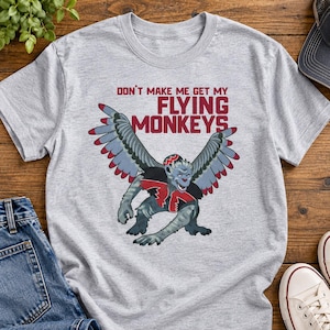 Pode incluir: Uma t-shirt cinzenta mesclada de manga curta com o gráfico de um macaco alado a usar um colete vermelho e preto. Acima do macaco, o texto "DON'T MAKE ME GET MY FLYING MONKEYS" está escrito em letras maiúsculas vermelho-escuras.