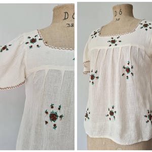 May include: A cream-colored blouse with a square neckline and short, flared sleeves. The garment features small, embroidered floral motifs in red and green scattered across the front and sleeves, with decorative brown stitching along the neckline.