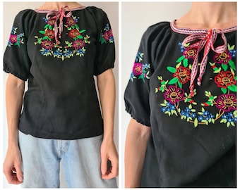 May include: Black embroidered peasant blouse with short, puffed sleeves. The blouse has a round neckline with a red and white ribbon tie. The floral embroidery features red, pink, blue, and green colours. The blouse is a vintage style.