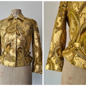 Puede incluir: Una chaqueta de tono dorado con un estampado de cachemira en tonos verde oliva, marrón y crema. La chaqueta tiene cuello, mangas largas y un cinturón que ajusta la cintura con un detalle de hebilla en el lateral, expuesta sobre un maniquí.