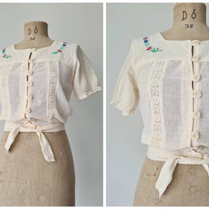 May include: A cream-colored short-sleeve blouse featuring a square neckline, a front button closure, and a tie-waist detail. The garment includes vertical lace panels and small embroidered floral accents in pink, blue, and green near the shoulders.