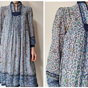 Könnte beinhalten: Ein langärmliges Kleid mit einem Blumenmuster in Blau-, Grün- und Cremetönen. Das Kleid hat einen hohen Ausschnitt mit dunkelblauer Bordüre und einem dekorativen Einsatz. Die Ärmel und der Saum sind ebenfalls dunkelblau eingefasst.