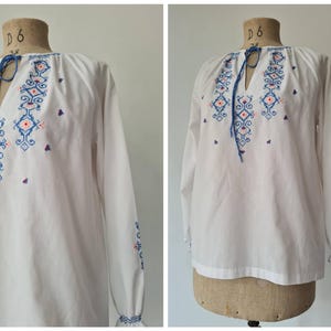 May include: A white long-sleeve blouse featuring a blue and orange geometric cross-stitch pattern on the chest and sleeves. The garment has a split neckline with a blue braided tie closure and ruffled cuffs, displayed on a dress form mannequin.