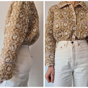 May include: A brown and white floral quilted jacket with a collar and button closure. The jacket features red and white trim and long sleeves. It is worn with white jeans. The jacket's design is reminiscent of vintage styles.