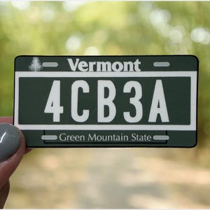 May include: A rectangular Vermont license plate sticker featuring a dark green background with white text. The top reads Vermont with a small tree icon, the center displays the alphanumeric code 4CB3A, and the bottom reads Green Mountain State.