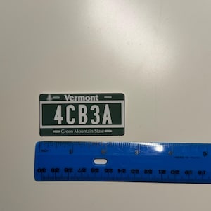May include: A rectangular Vermont license plate replica in dark green and white, featuring the text Vermont, 4CB3A, and Green Mountain State. A blue plastic ruler is positioned below the plate, showing it measures approximately 3.5 inches in width.